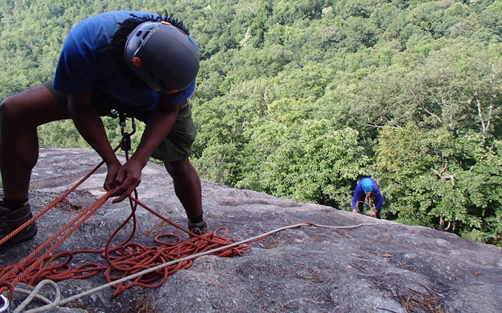 In the foreground, one person wearing safety gear is secured by ropes as they look down a rocky slope to another person, also wearing safety gear and secured by ropes, who is making their way up.